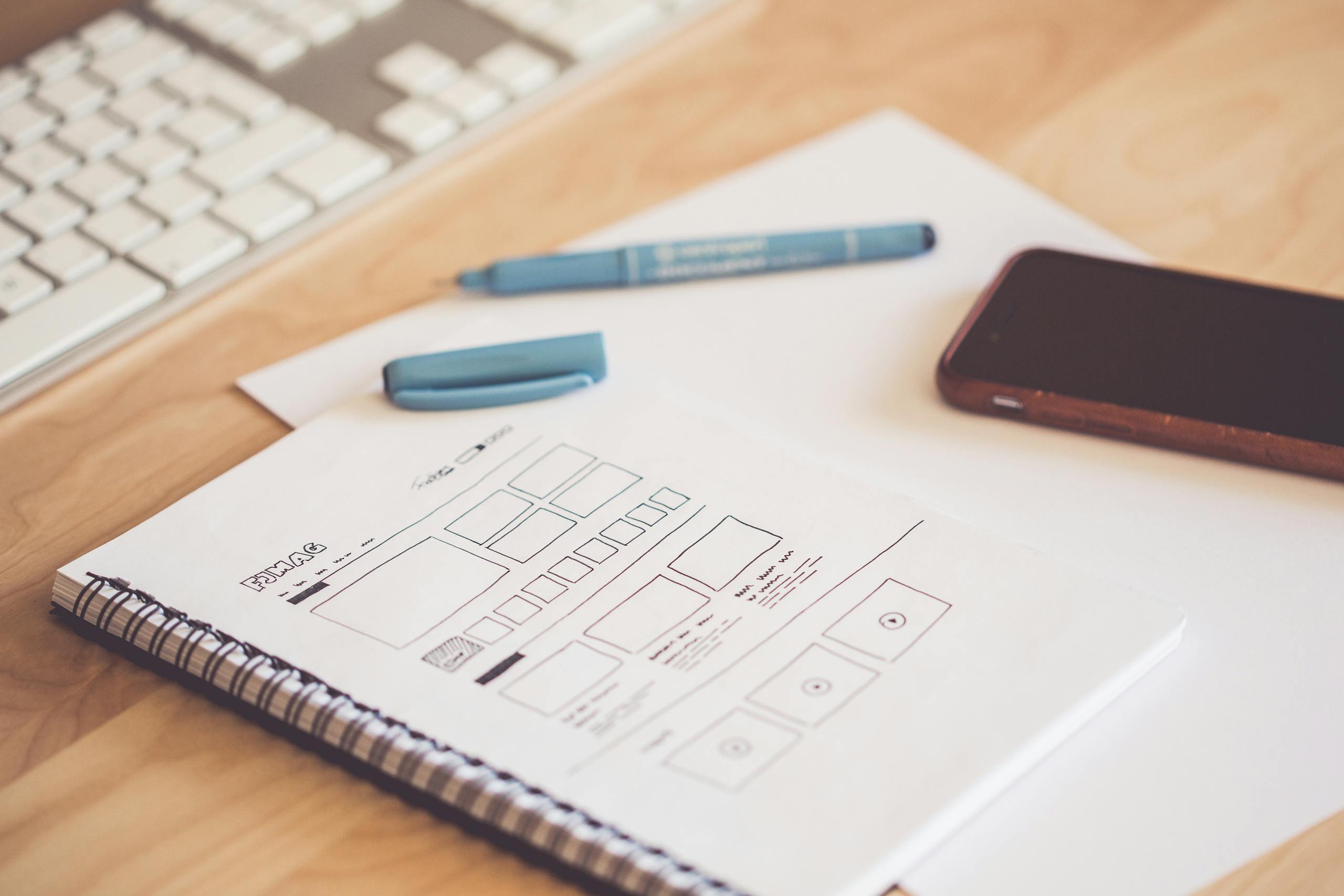 A neat workspace featuring a sketchpad with wireframes, a smartphone, and a keyboard on a wooden desk. website and social media services for small businesses | Oaken Co